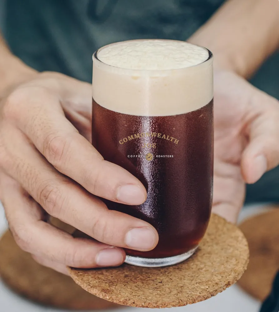 Hands holding a Commonwealth Joe glass of cold brew coffee with a thick foam head, resting on a cork coaster