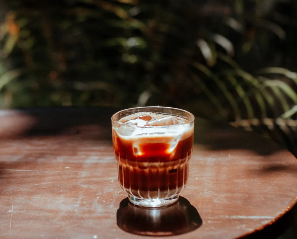A glass of cold brew coffee with a pale head on a table, palm foliage in warm daylight behind