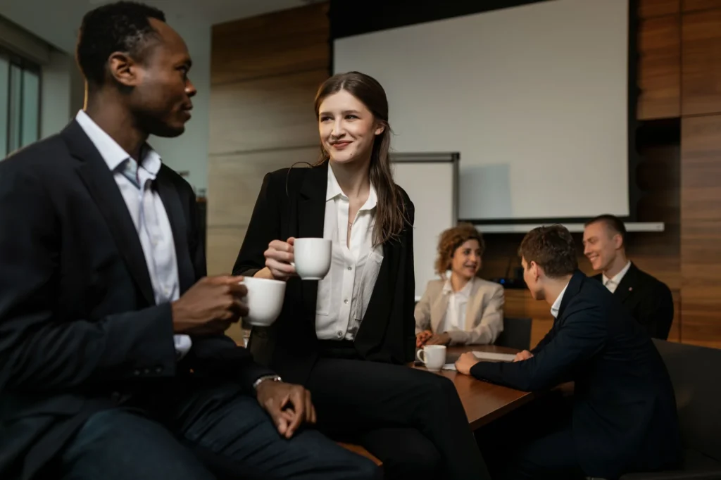 Colleagues talking over coffee in a meeting room while a project team works at the table behind