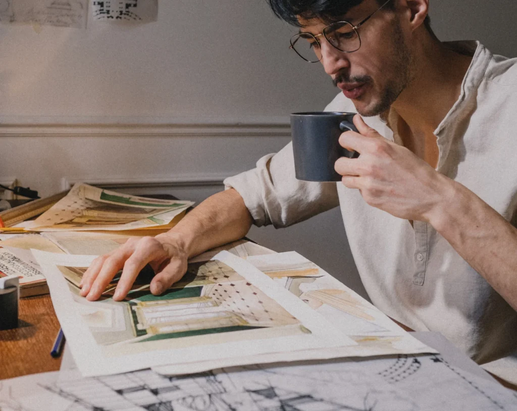 An engineer drinking coffee at a desk covered with technical drawings and elevations