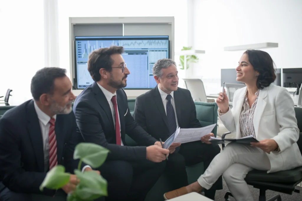 A team of finance professionals reviewing reports in front of a wall monitor showing market charts