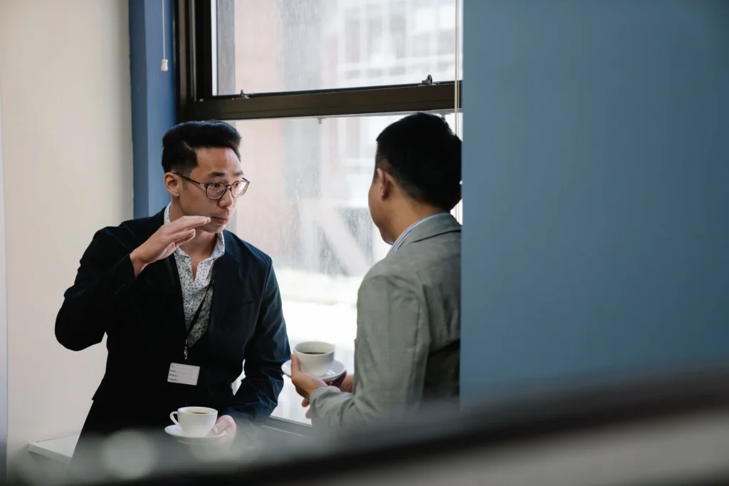 Two colleagues holding espresso cups during a conversation beside an office window