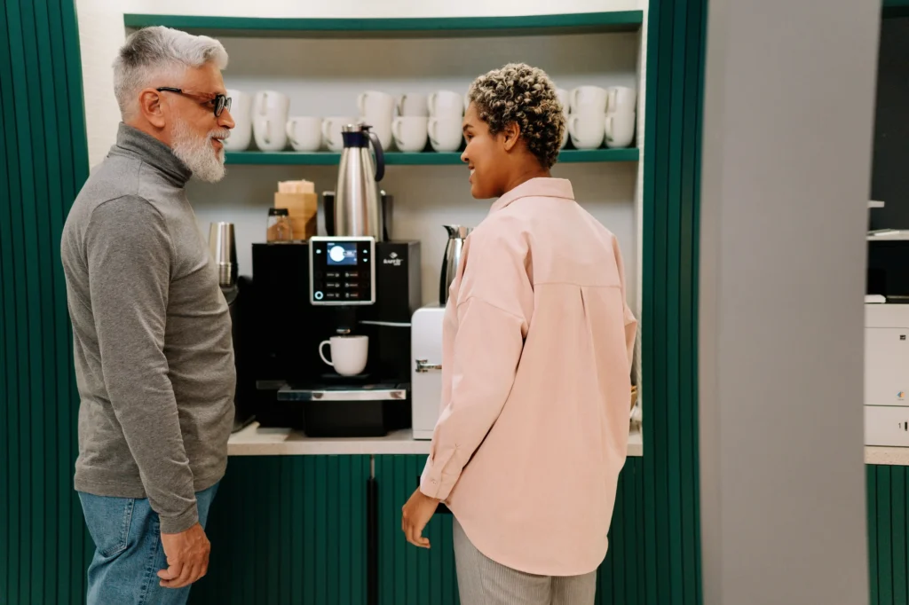 Two colleagues talking at an office coffee station with a bean-to-cup machine, carafes and a shelf of cups