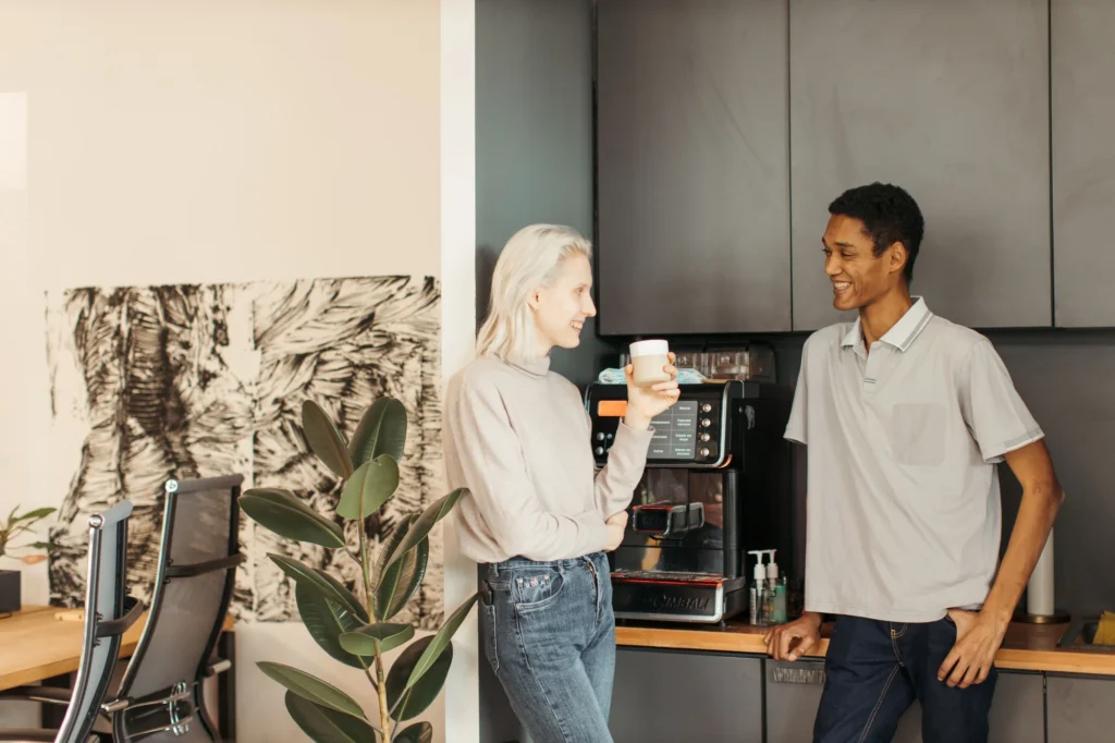 Two colleagues at an office kitchen counter, one holding a fresh cup beside a bean-to-cup coffee machine