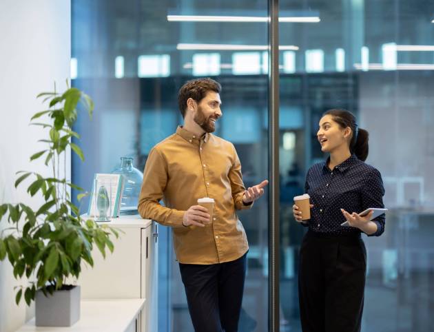Two coworkers engage in a friendly conversation while holding coffee cups in a bright office.